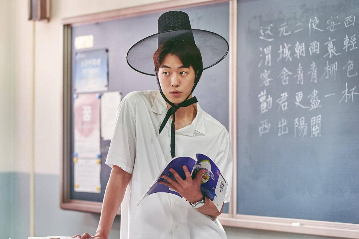Seorang pria mengenakan topi tradisional Korea dan memegang buku di ruang kelas dengan papan tulis berisi tulisan aksara Korea di belakangnya.