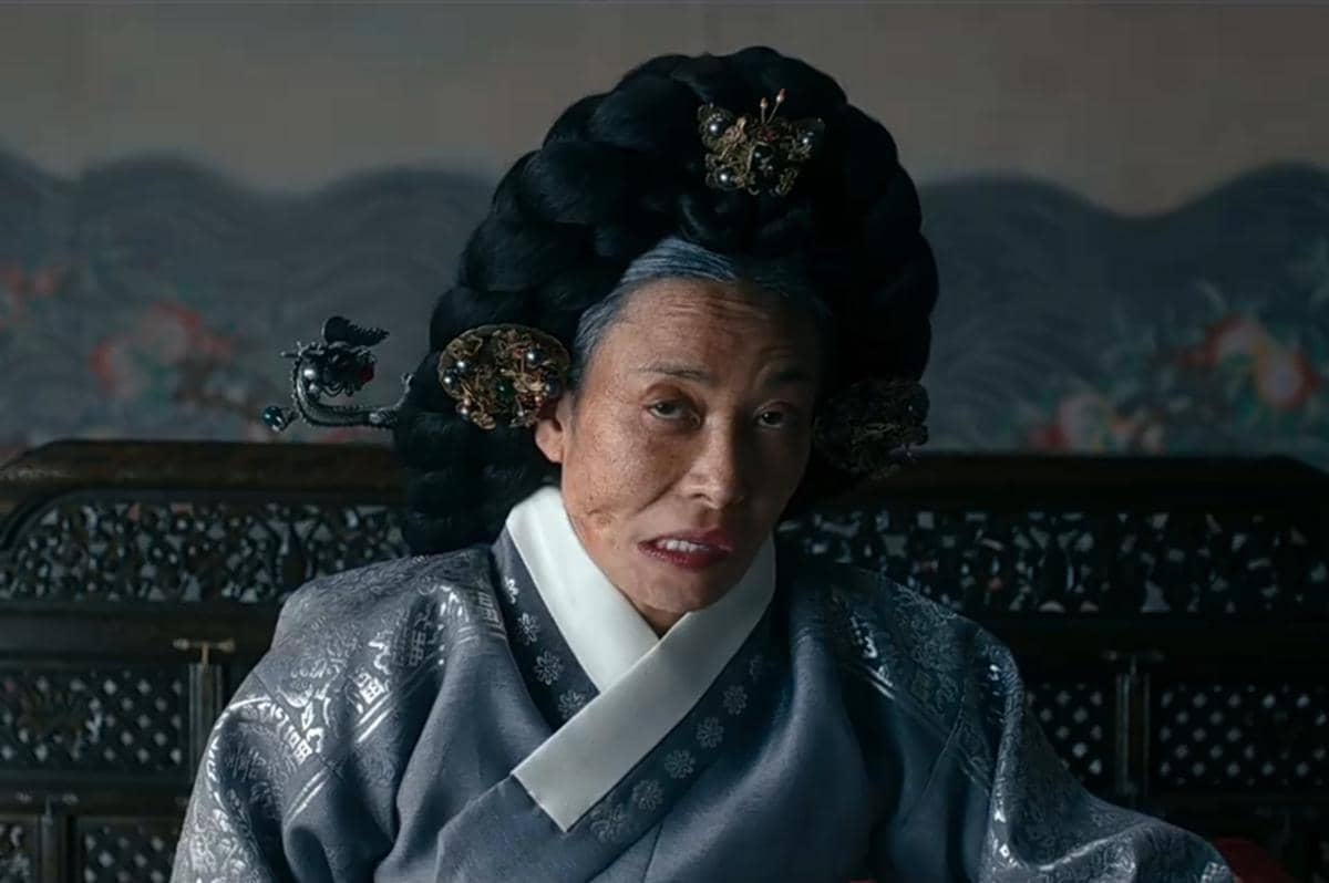 Seorang karakter mengenakan hanbok berwarna abu-abu dengan tatanan rambut tradisional besar dan hiasan kepala rumit dalam adegan The East Palace.