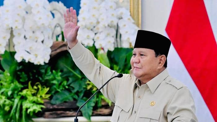 Prabowo Subianto mengenakan kemeja krem dan peci hitam melambaikan tangan kanan di depan mikrofon dan bendera Merah Putih.
