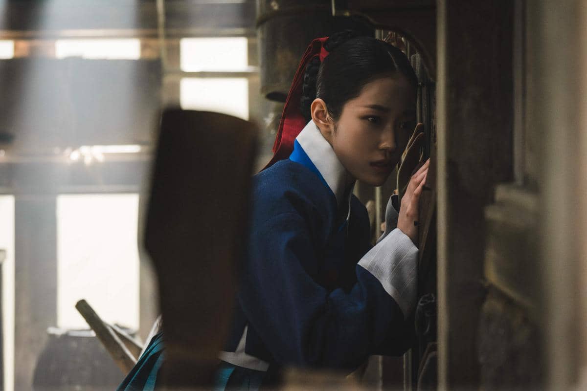Seorang perempuan mengenakan hanbok biru dengan pita merah di rambutnya tampak mengintip melalui celah dinding kayu dalam suasana redup.