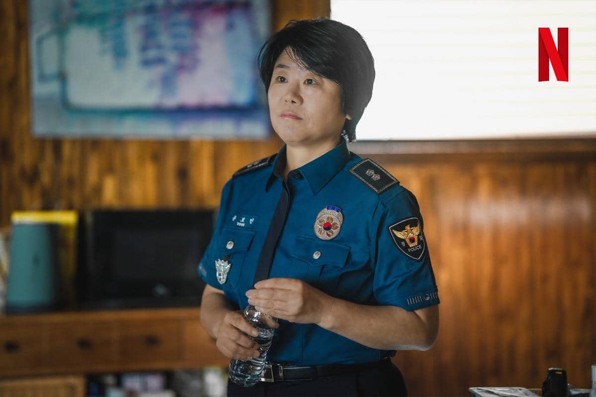 Seorang perempuan berseragam polisi berdiri di dalam ruangan kayu sambil memegang botol air, dalam cuplikan drama The Frog di Netflix.