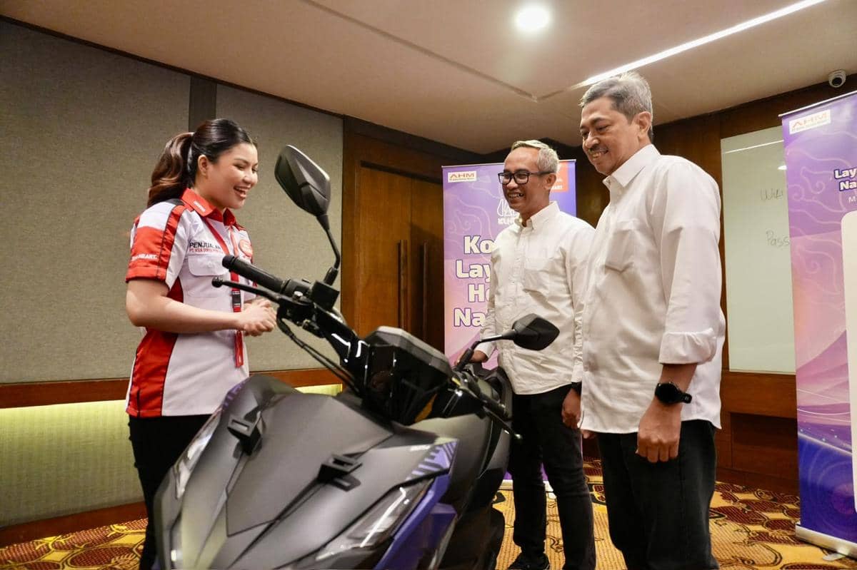 Seorang peserta berseragam AHM berbincang dengan dua pria di depan sepeda motor dalam acara kompetisi layanan nasional.