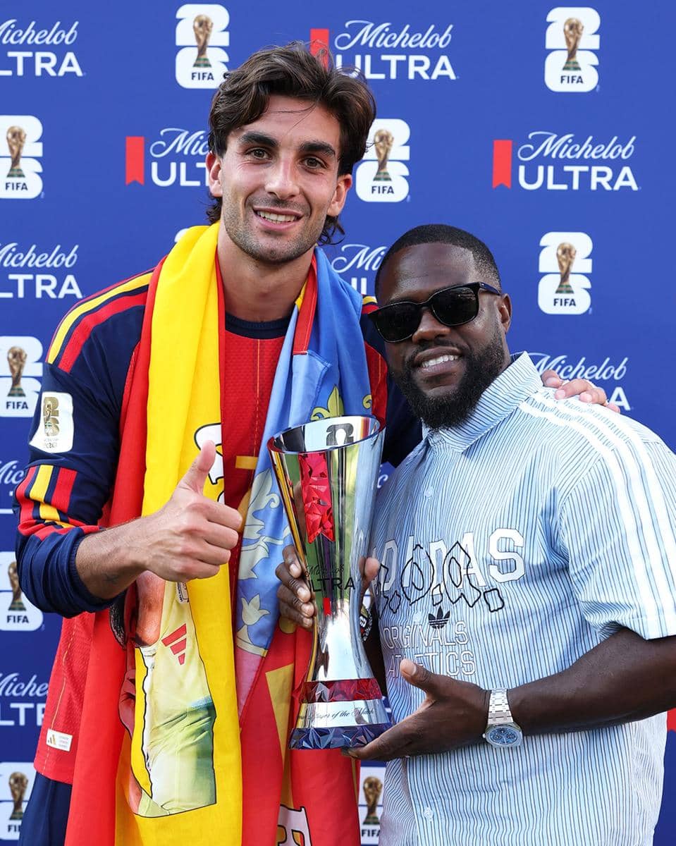 Dua pria berpose sambil memegang trofi di acara final Piala Dunia 2026 dengan latar belakang bertema FIFA dan sponsor Michelob Ultra.