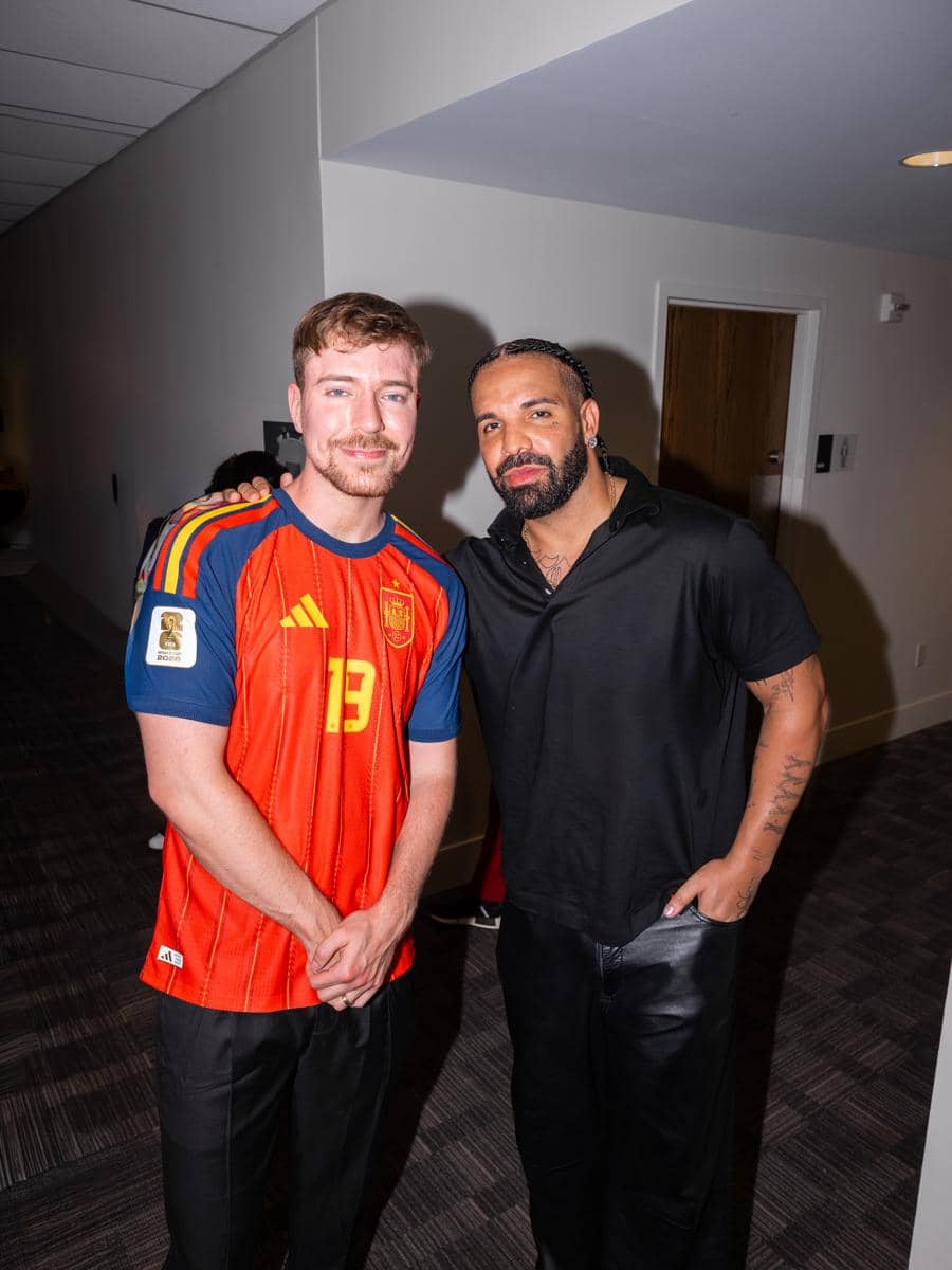 Dua pria berpose bersama di dalam ruangan, salah satunya mengenakan jersey sepak bola Spanyol dan yang lain mengenakan kemeja hitam.