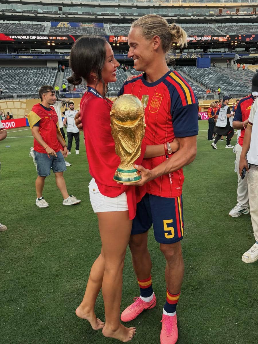 Seorang pemain tim nasional Spanyol memegang trofi Piala Dunia sambil berpelukan dengan pasangannya di lapangan stadion.