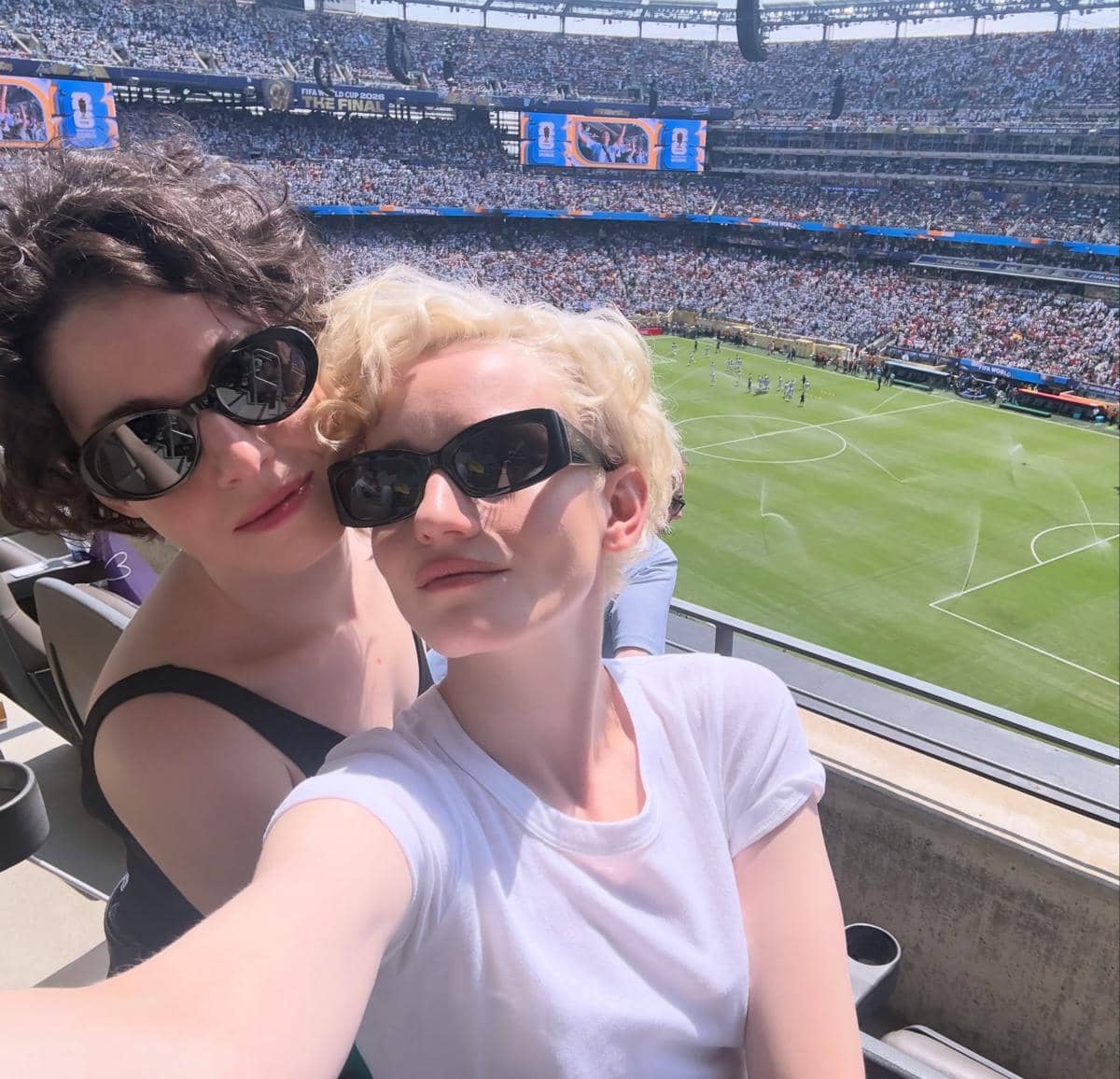 Dua orang berpose selfie di tribun stadion saat final Piala Dunia 2026 dengan latar belakang lapangan dan penonton penuh.