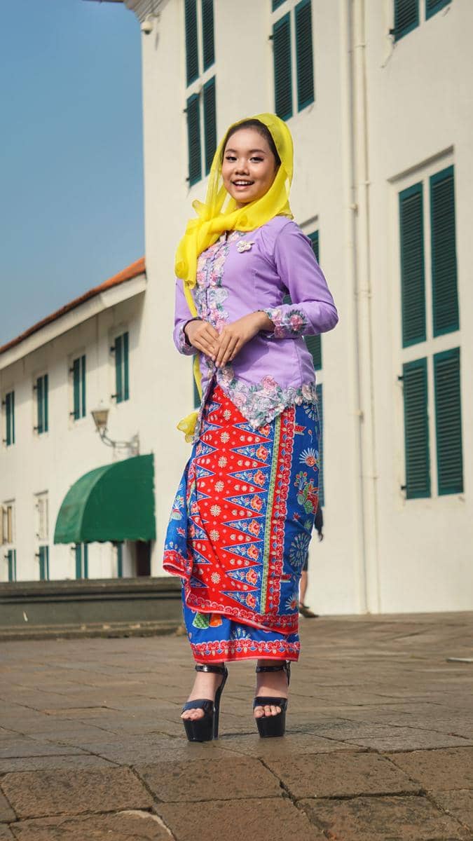 Perempuan mengenakan kebaya encim ungu dengan selendang kuning dan kain batik merah biru berdiri di area berarsitektur kolonial.