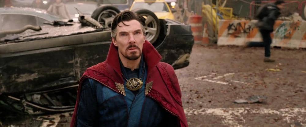Doctor Strange mengenakan jubah merah dan pakaian biru berdiri di jalan dengan mobil terbalik di belakangnya dalam adegan film Marvel.
