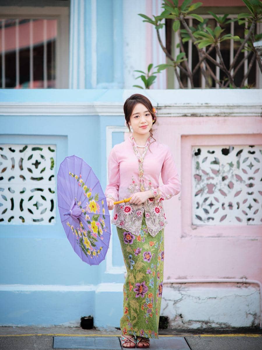 Perempuan mengenakan kebaya encim warna lembut dengan kain batik hijau bermotif bunga sambil memegang payung ungu bermotif floral.