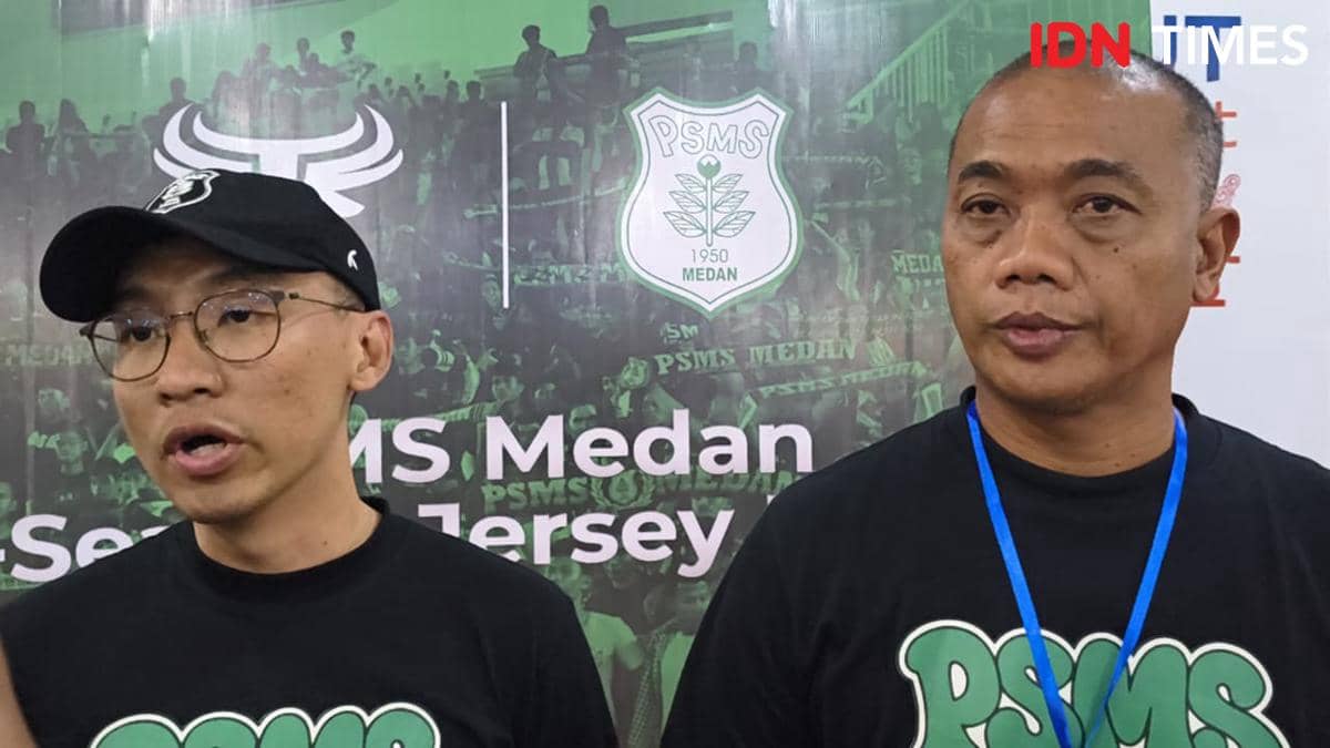 Dua pria mengenakan kaus hitam bertuliskan PSMS berdiri di depan latar bertema PSMS Medan dengan logo klub berwarna hijau putih.