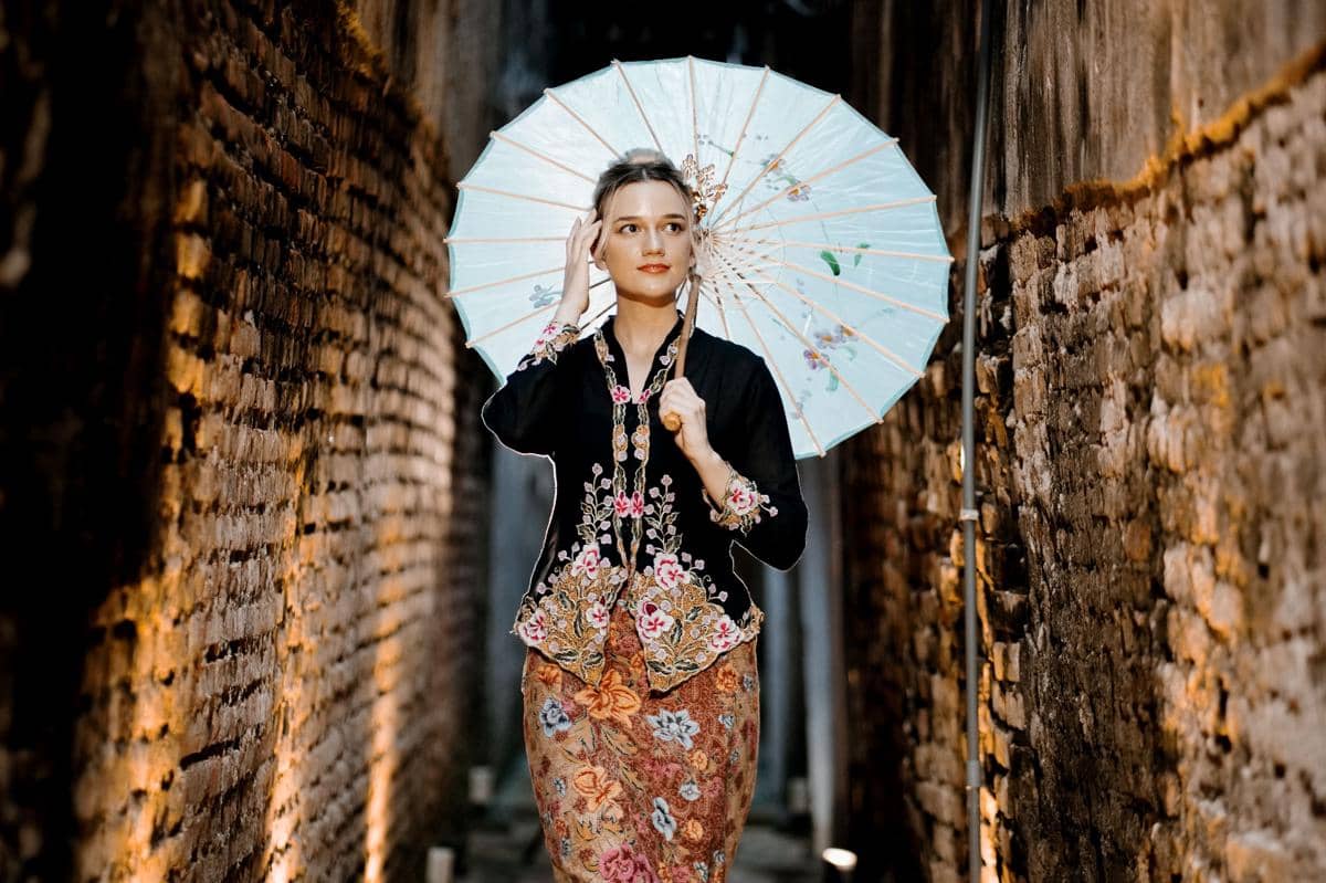 Perempuan mengenakan kebaya encim warna gelap bermotif bunga dengan kain batik, memegang payung kertas di lorong berdinding bata.