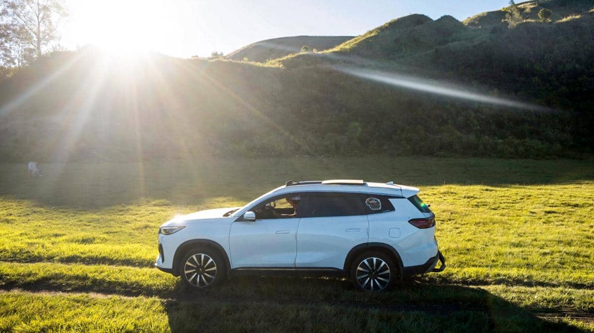 Sebuah mobil SUV putih Chery melintas di area perbukitan hijau dengan sinar matahari pagi yang terang di latar belakang.