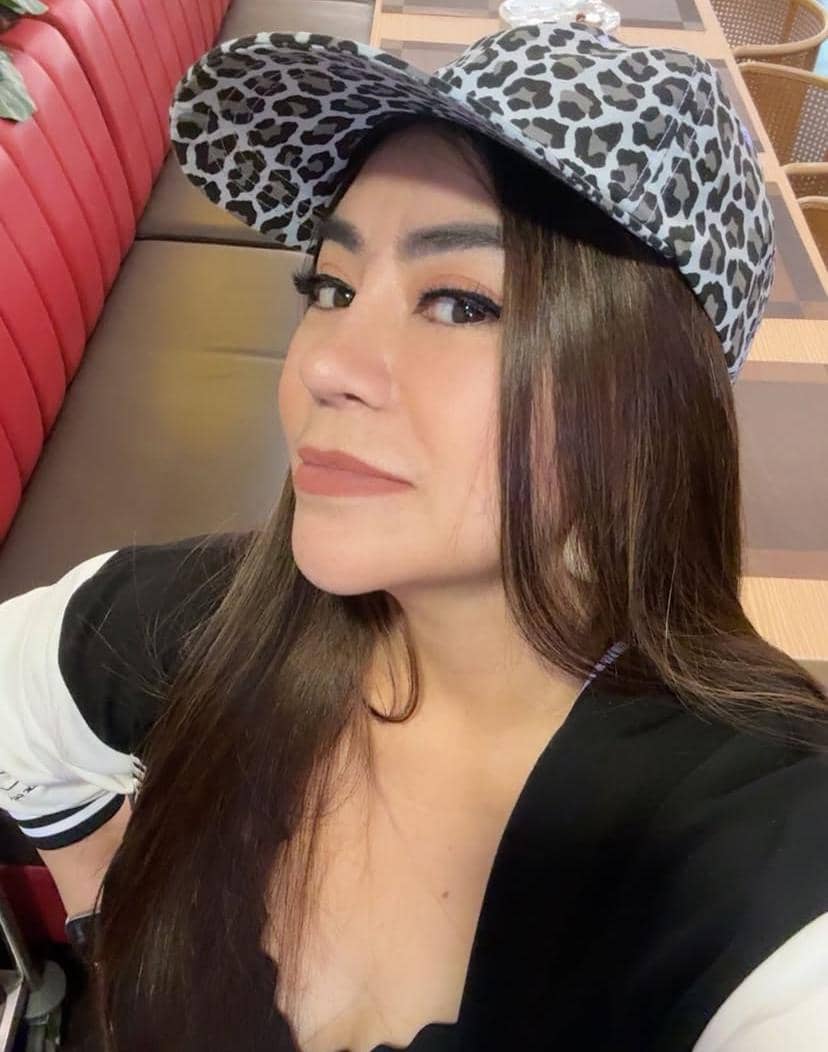 Seorang perempuan berambut panjang mengenakan topi bermotif leopard dan pakaian hitam putih sedang berpose di tempat duduk restoran.