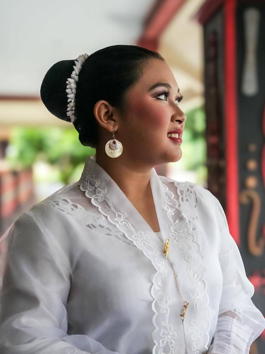 Perempuan mengenakan kebaya encim putih dengan sanggul rapi dan anting bundar, tersenyum sambil menatap ke samping di area terbuka.