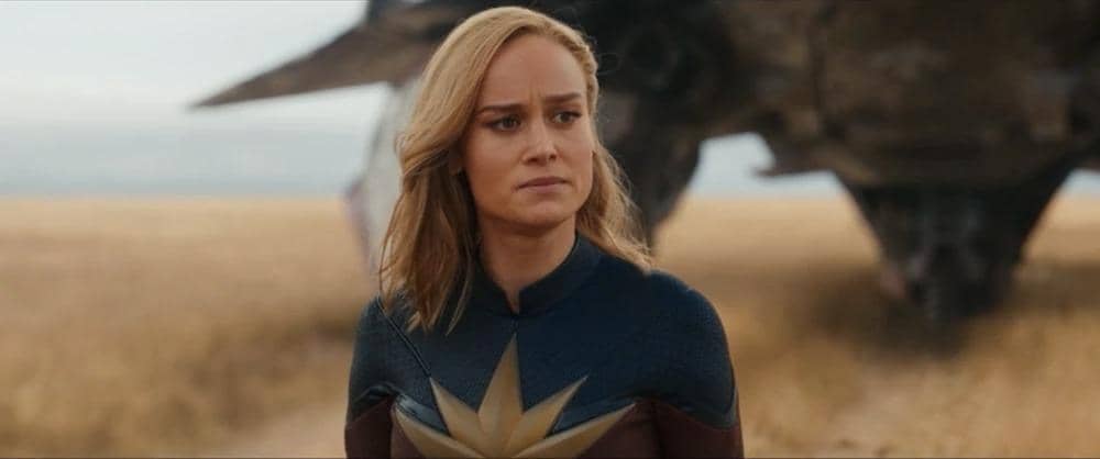 Captain Marvel berdiri di padang rumput dengan ekspresi serius mengenakan kostum biru dan emas, dengan pesawat luar angkasa di latar belakang.