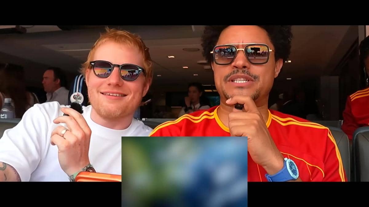 Dua pria mengenakan kacamata hitam duduk di tribun menonton final Piala Dunia 2026, salah satunya memakai kaus merah bergaris kuning.