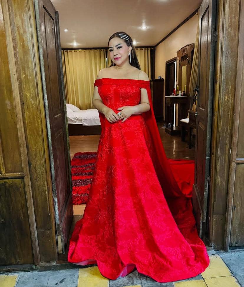 Seorang wanita mengenakan gaun merah panjang tanpa lengan berdiri di antara dua pintu kayu di dalam ruangan bergaya klasik.