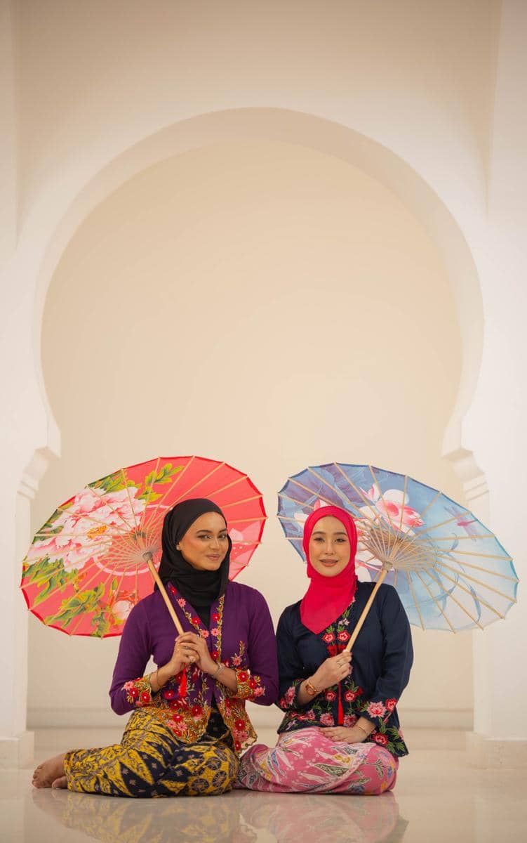 Dua wanita mengenakan kebaya encim bordir floral berwarna cerah sambil memegang payung hias bermotif bunga di ruang bercorak arsitektur klasik.