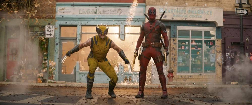 Dua karakter superhero, Deadpool dan Wolverine, berdiri di jalan dengan latar bangunan toko yang rusak dan asap di sekitar mereka.