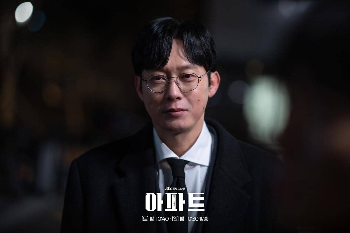 Seorang pria berkacamata mengenakan jas hitam dan dasi berdiri di malam hari dalam cuplikan drama The Apartment Job.