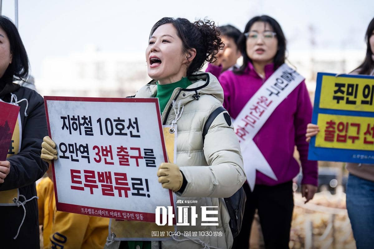 Seorang wanita memegang papan protes bertuliskan huruf Korea sambil berteriak di tengah kelompok orang dalam cuplikan drama The Apartment Job.