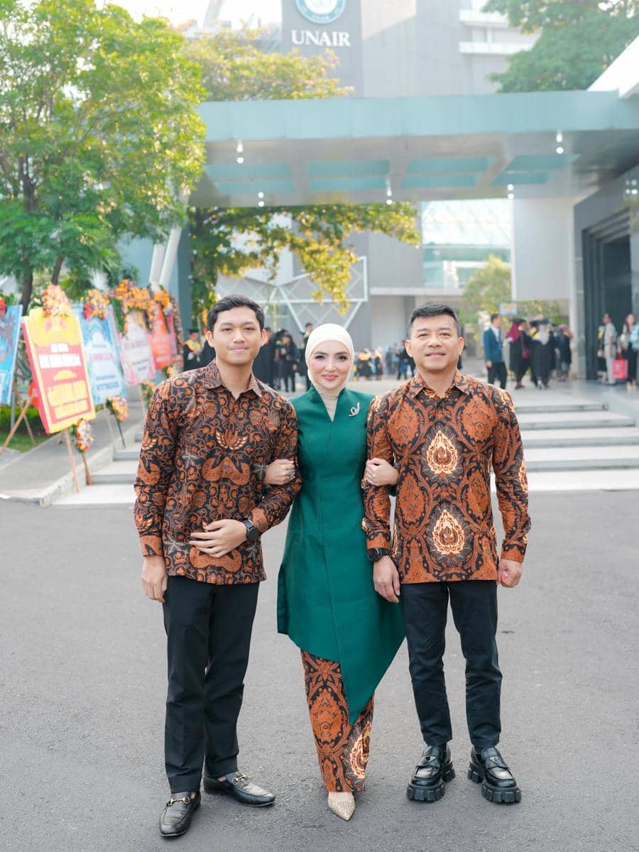 Seorang wanita berhijab berbusana hijau berpose bersama dua pria mengenakan batik cokelat di depan gedung Universitas Airlangga.