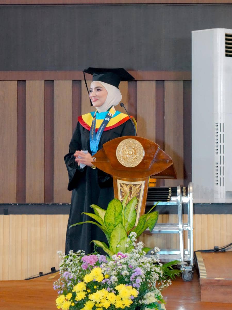 Seorang perempuan berhijab mengenakan toga wisuda berdiri di podium dengan latar ruang auditorium dan hiasan bunga di depannya.