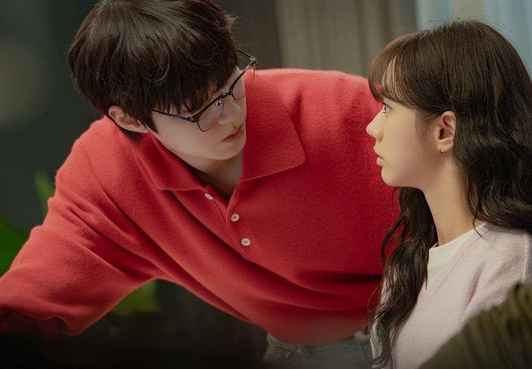 Dua karakter dalam drama Korea Dream to You saling menatap intens, pria berkacamata mengenakan sweater merah dan wanita berambut panjang duduk di depannya.