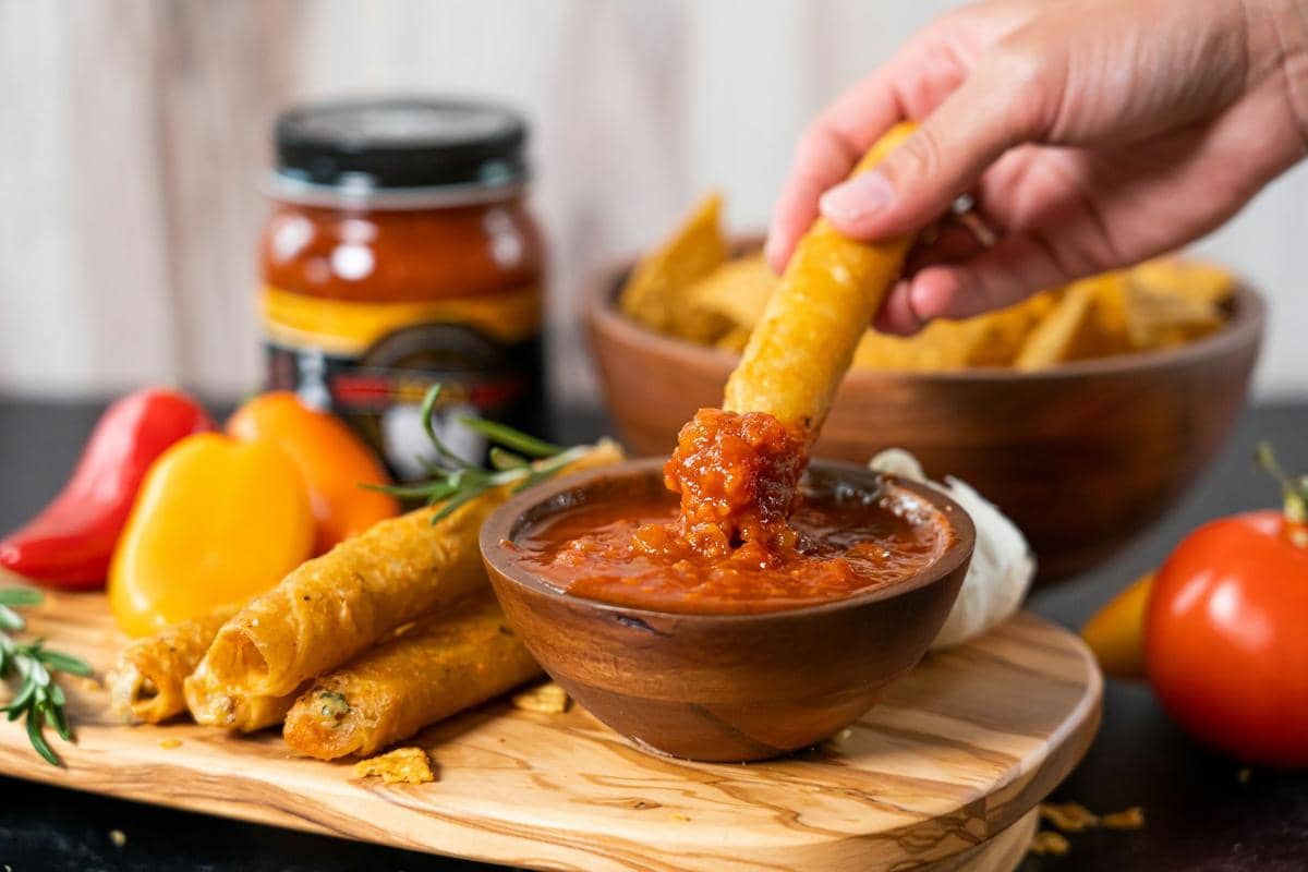 Tangan mencelupkan taquito ke dalam mangkuk salsa taquera merah dengan latar belakang cabai dan sayuran segar di atas meja kayu.