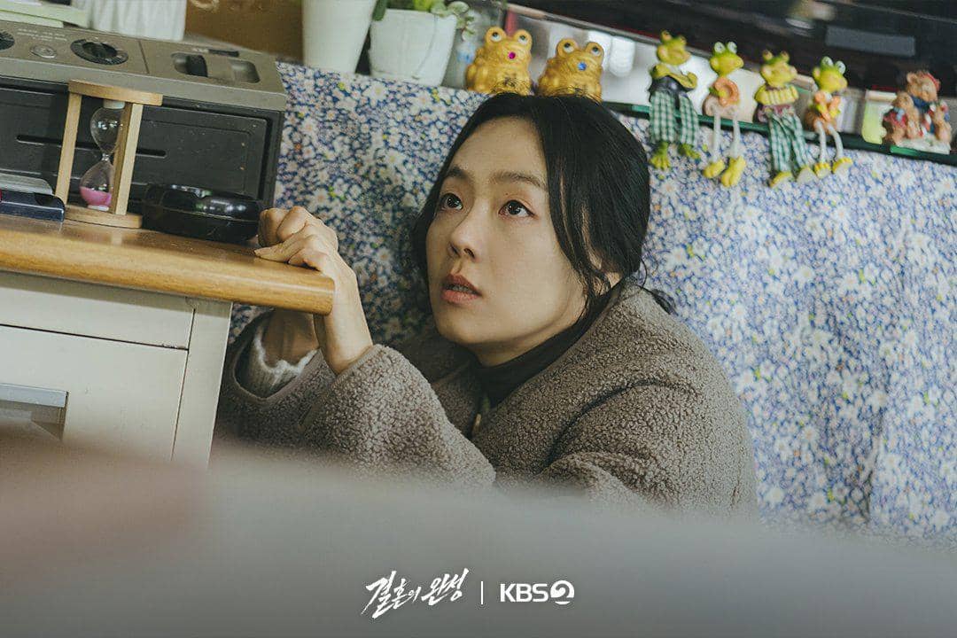 Seorang wanita berjaket tebal bersembunyi di bawah meja dengan ekspresi tegang di adegan drama The Husband produksi KBS.