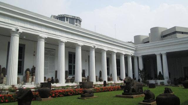 Bagian halaman Museum Nasional Indonesia dengan deretan pilar putih klasik dan koleksi arca batu di area taman hijau.