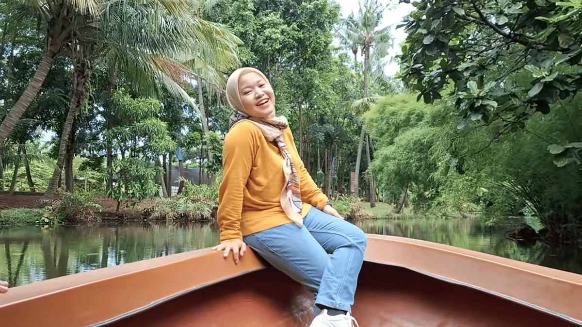 Seorang perempuan duduk santai di atas perahu yang mengapung di area hijau Fauna Land Ancol dengan latar pepohonan rindang.