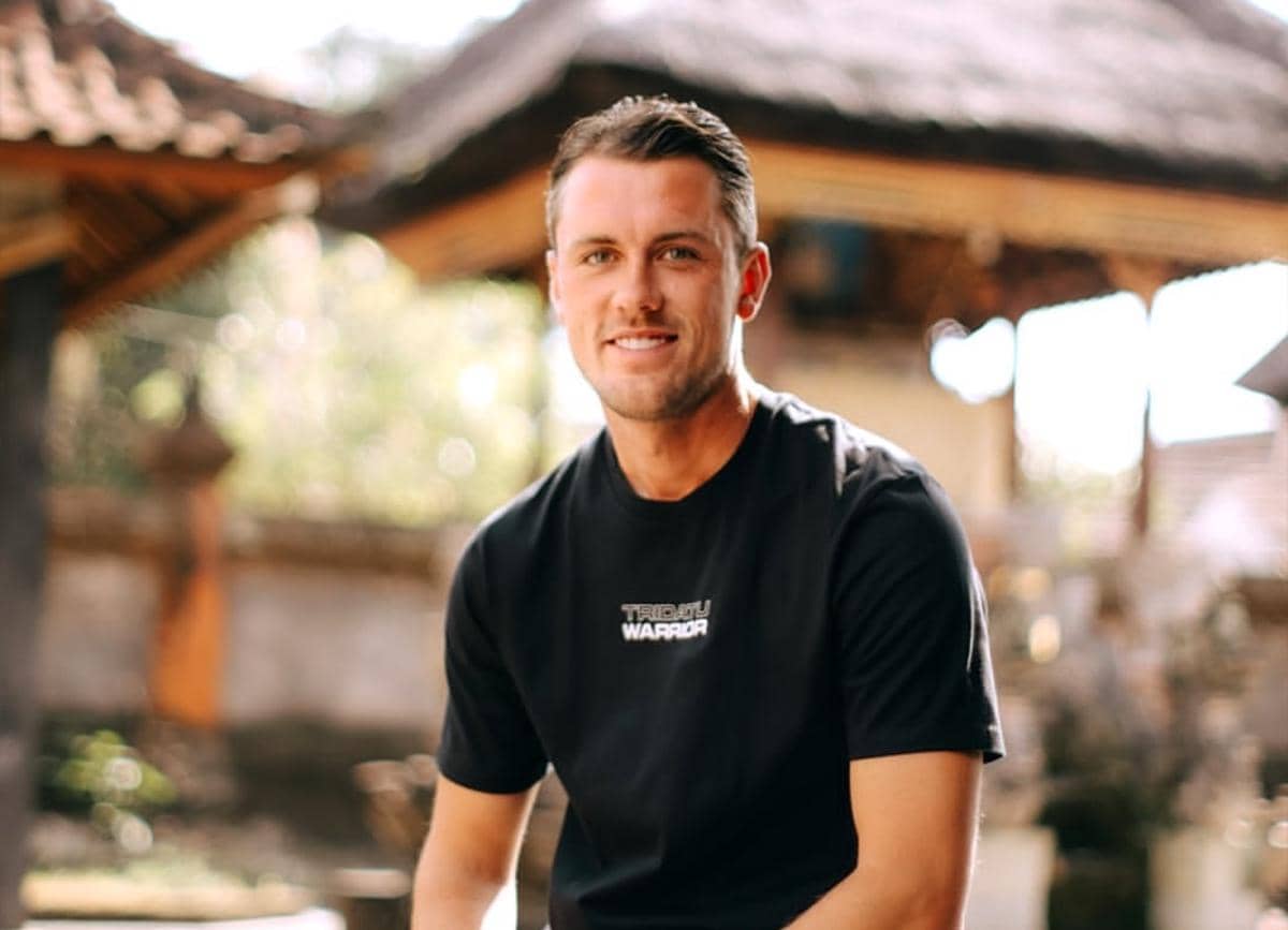 Seorang pria mengenakan kaus hitam duduk santai di area luar ruangan dengan latar bangunan bergaya tradisional Bali.