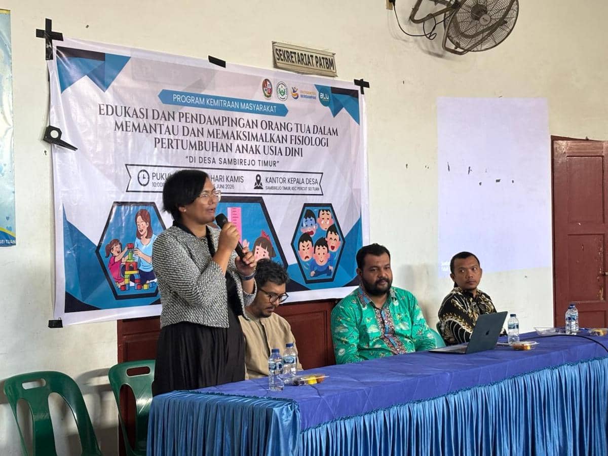 Seorang pembicara dari FIK Unimed memberikan sosialisasi di Desa Sambirejo Timur dengan beberapa peserta duduk di meja berlapis kain biru.