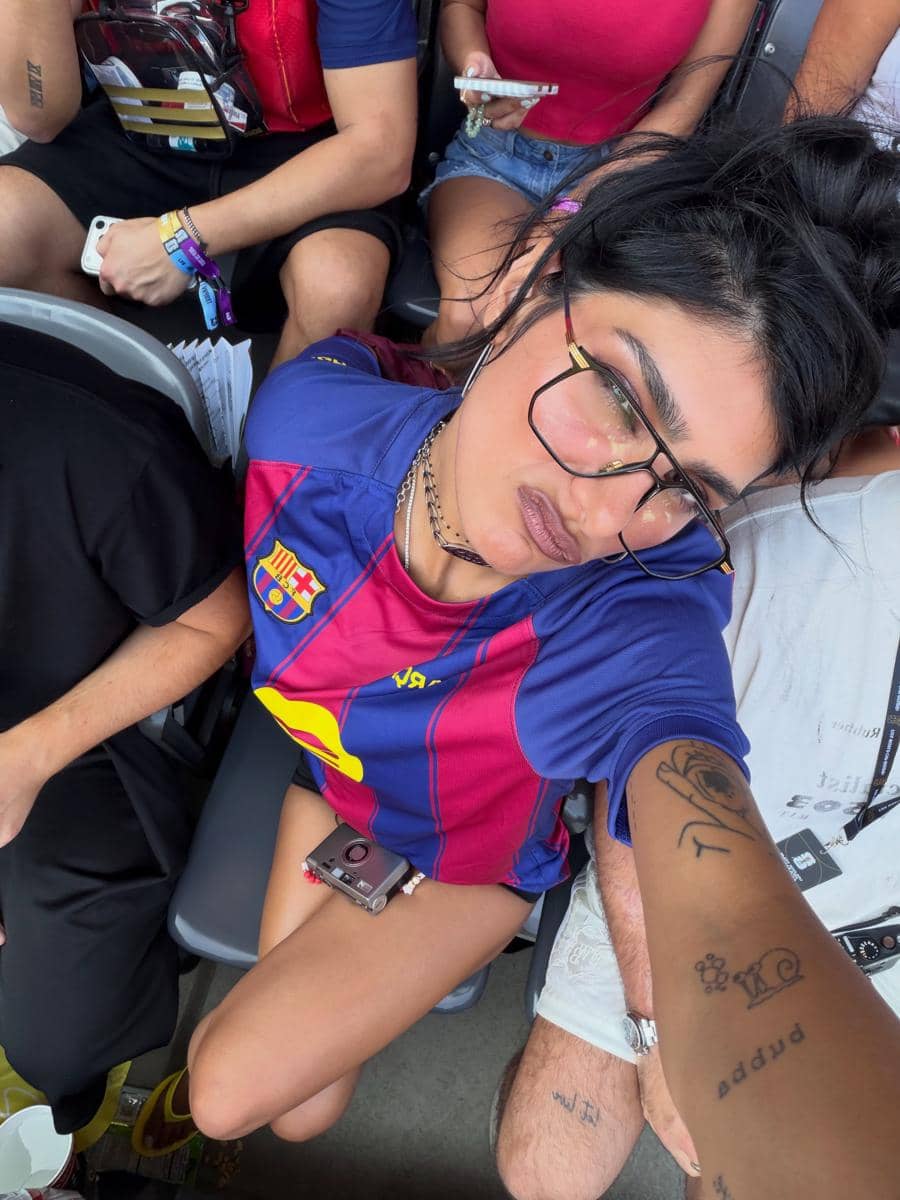 Seorang perempuan mengenakan jersey sepak bola warna biru dan merah duduk di tribun stadion bersama penonton lain sambil berfoto selfie.
