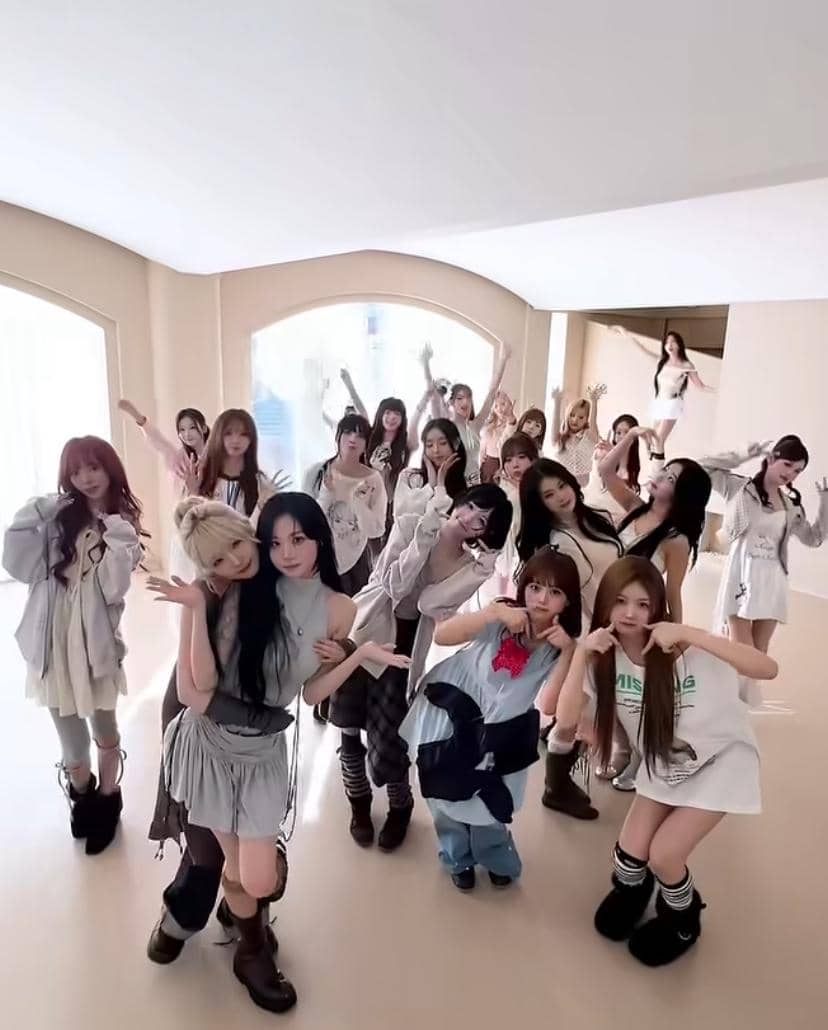 Sekelompok anggota girl group berdiri dan berpose bersama di dalam ruangan bercahaya alami dengan dinding berwarna krem.
