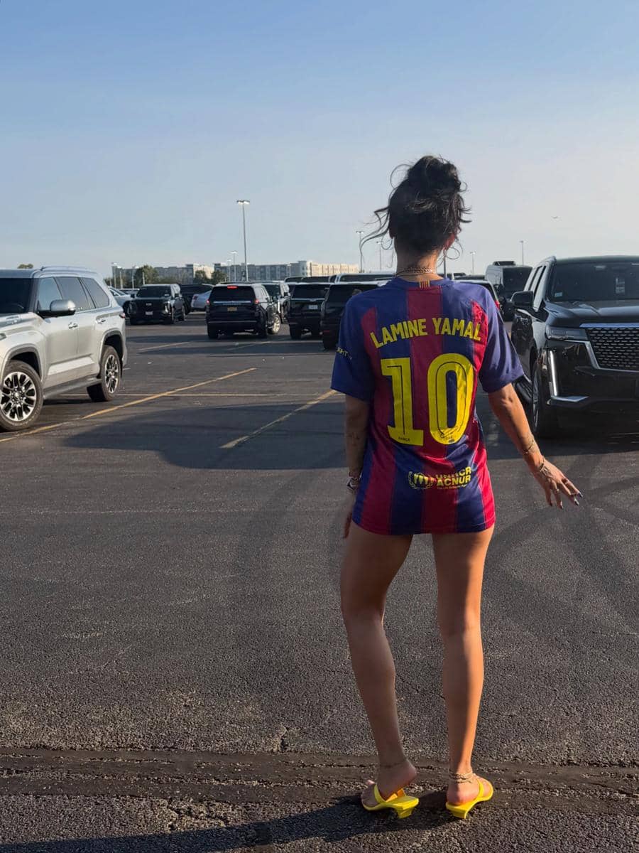 Seorang perempuan mengenakan jersey sepak bola bernomor 10 bertuliskan Lamine Yamal berdiri di area parkir dengan latar mobil dan langit cerah.