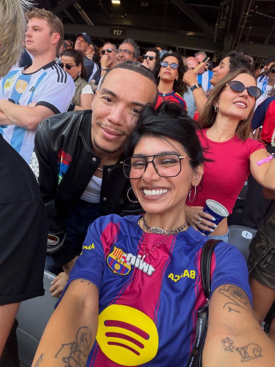 Seorang perempuan mengenakan jersey Barcelona berfoto selfie bersama seorang pria di tribun stadion yang dipenuhi penonton pertandingan sepak bola.