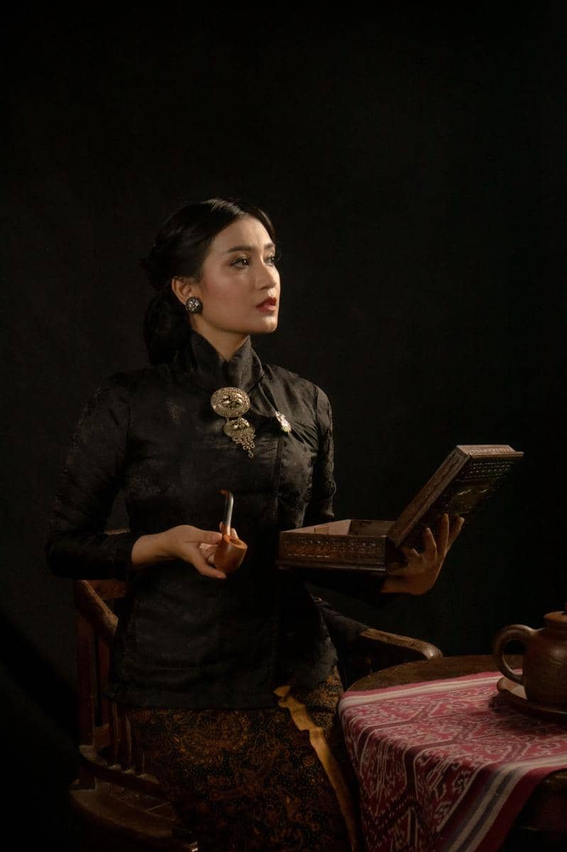 Perempuan mengenakan kebaya janggan hitam dengan bros perak, memegang kotak kayu dan pipa di ruangan bernuansa klasik.