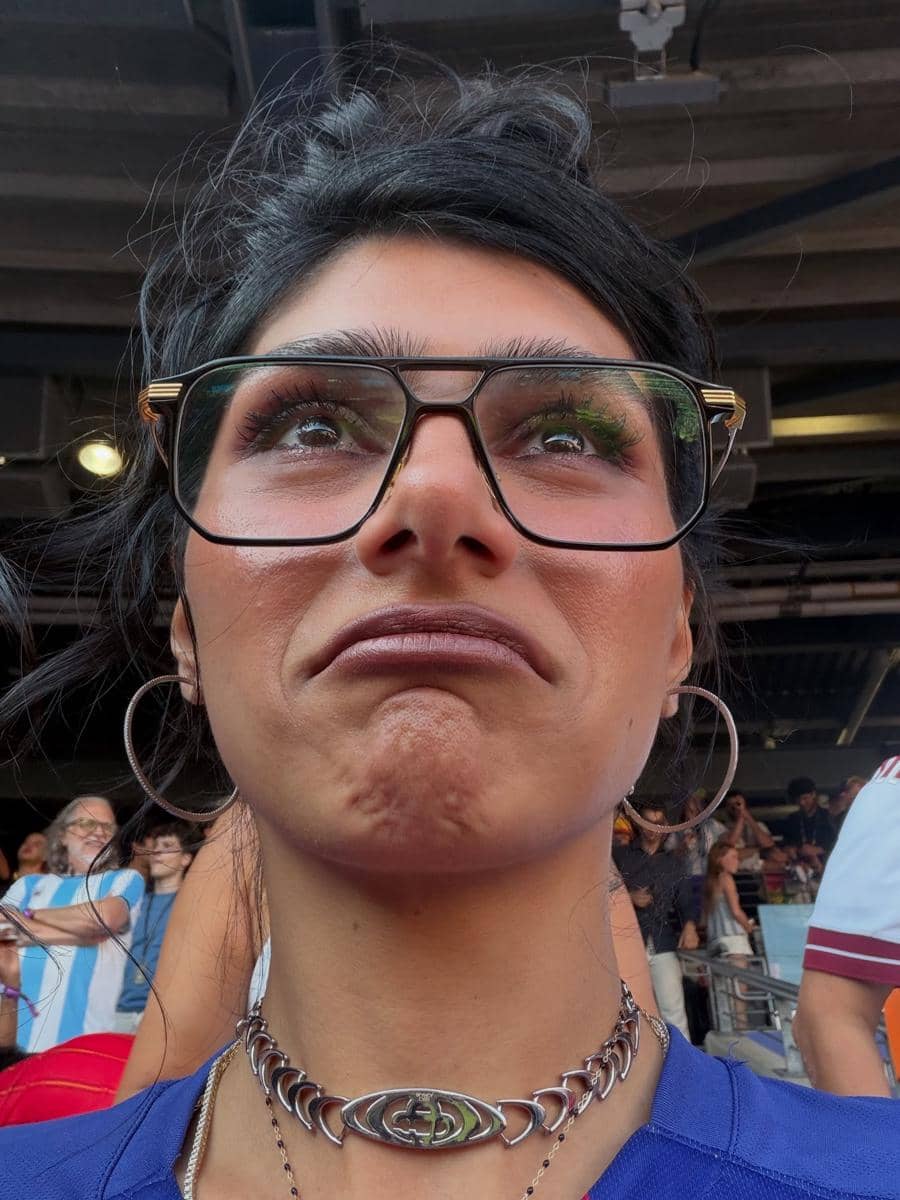 Seorang perempuan mengenakan kacamata besar dan kalung perak sedang berada di stadion dengan ekspresi wajah datar saat menonton pertandingan.