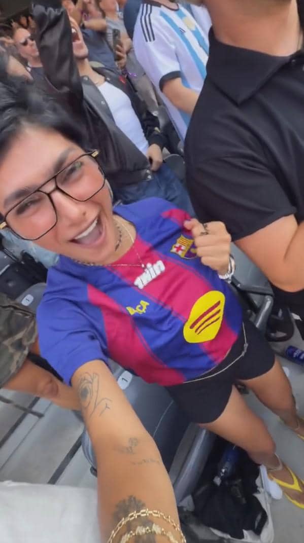 Seorang perempuan mengenakan kaus sepak bola biru-merah berpose gembira di stadion bersama penonton lain yang juga bersorak.