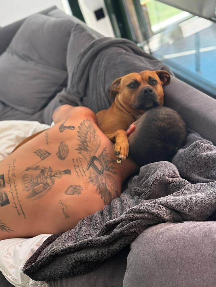 Seorang pria bertato berbaring di sofa sambil memeluk anjing berwarna cokelat, keduanya tampak santai di bawah selimut abu-abu.