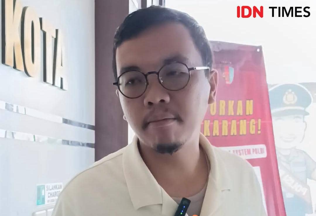 Seorang pria berkacamata mengenakan kemeja berkerah warna terang berdiri di dalam ruangan dengan latar tulisan dan poster berwarna merah.