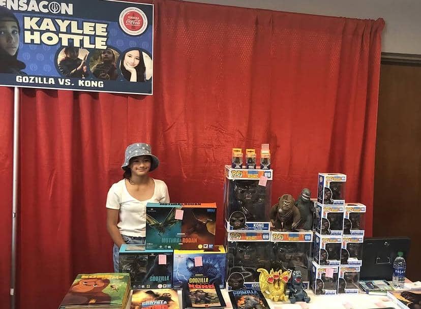 Seorang perempuan muda berdiri di belakang meja penuh mainan dan figur Godzilla vs. Kong di sebuah acara dengan latar tirai merah.