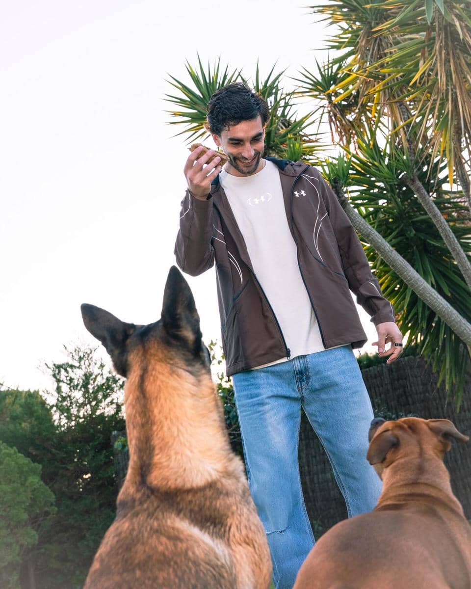 Seorang pria mengenakan jaket cokelat dan kaus putih sedang bermain dengan dua anjing di luar ruangan dengan latar pepohonan tropis.