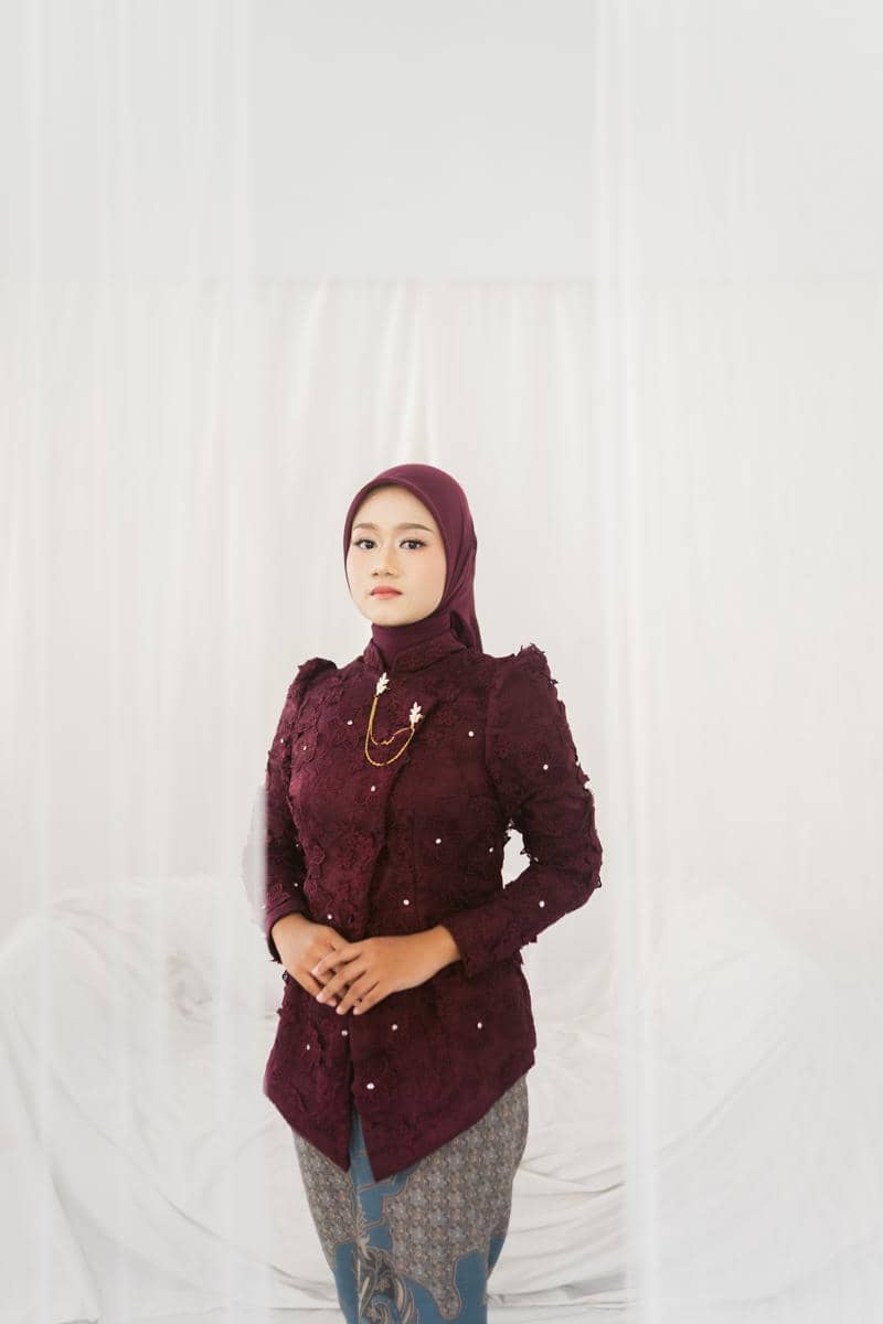 Perempuan mengenakan kebaya janggan berwarna merah marun dengan hijab senada dan kain batik bermotif di latar putih lembut.