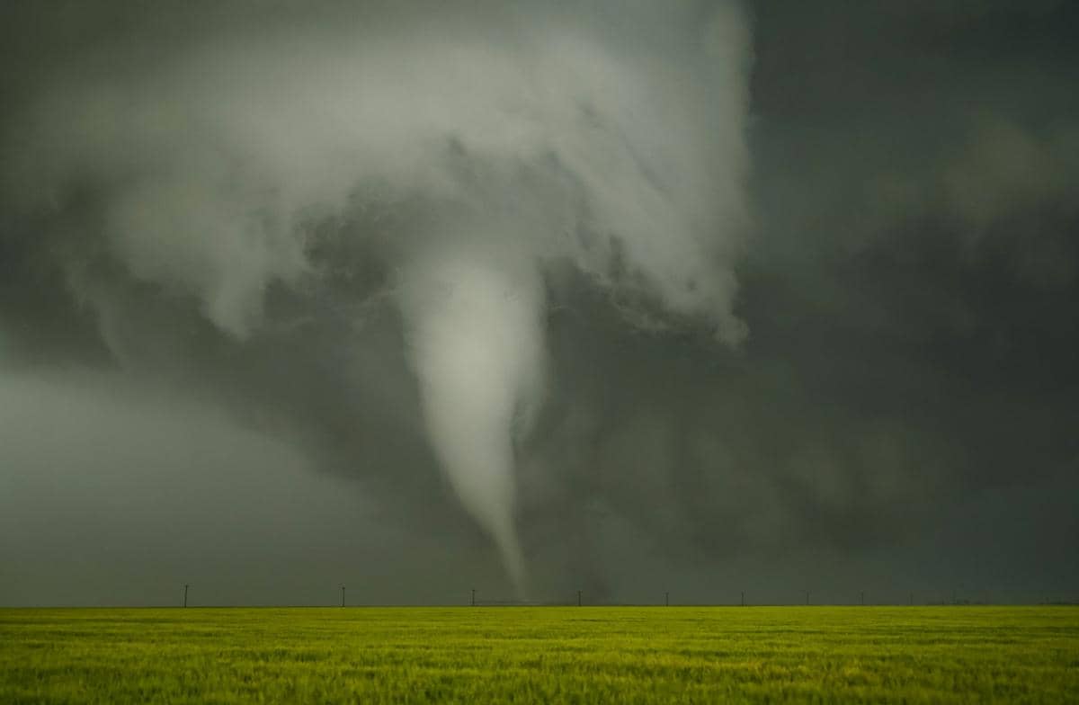 Tornado besar berputar di atas ladang hijau dengan langit gelap dan awan tebal, menandakan badai kuat di daerah terbuka.