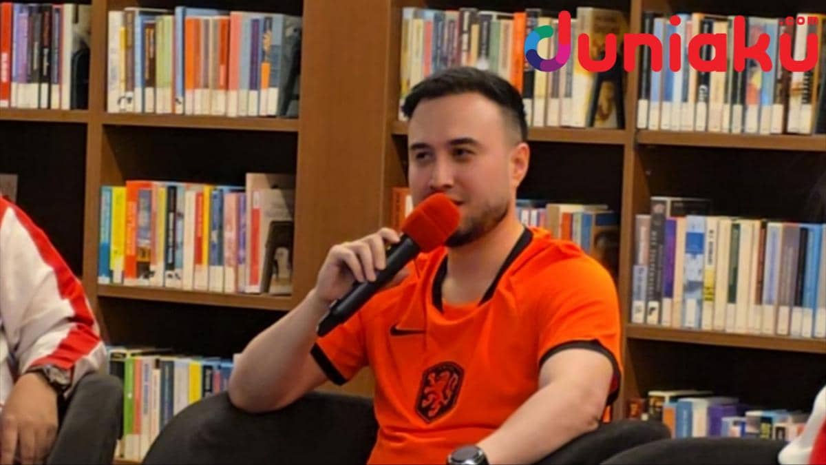 Seorang pria mengenakan jersey oranye sedang berbicara dengan mikrofon di depan rak buku selama panel diskusi di Erasmus Huis.