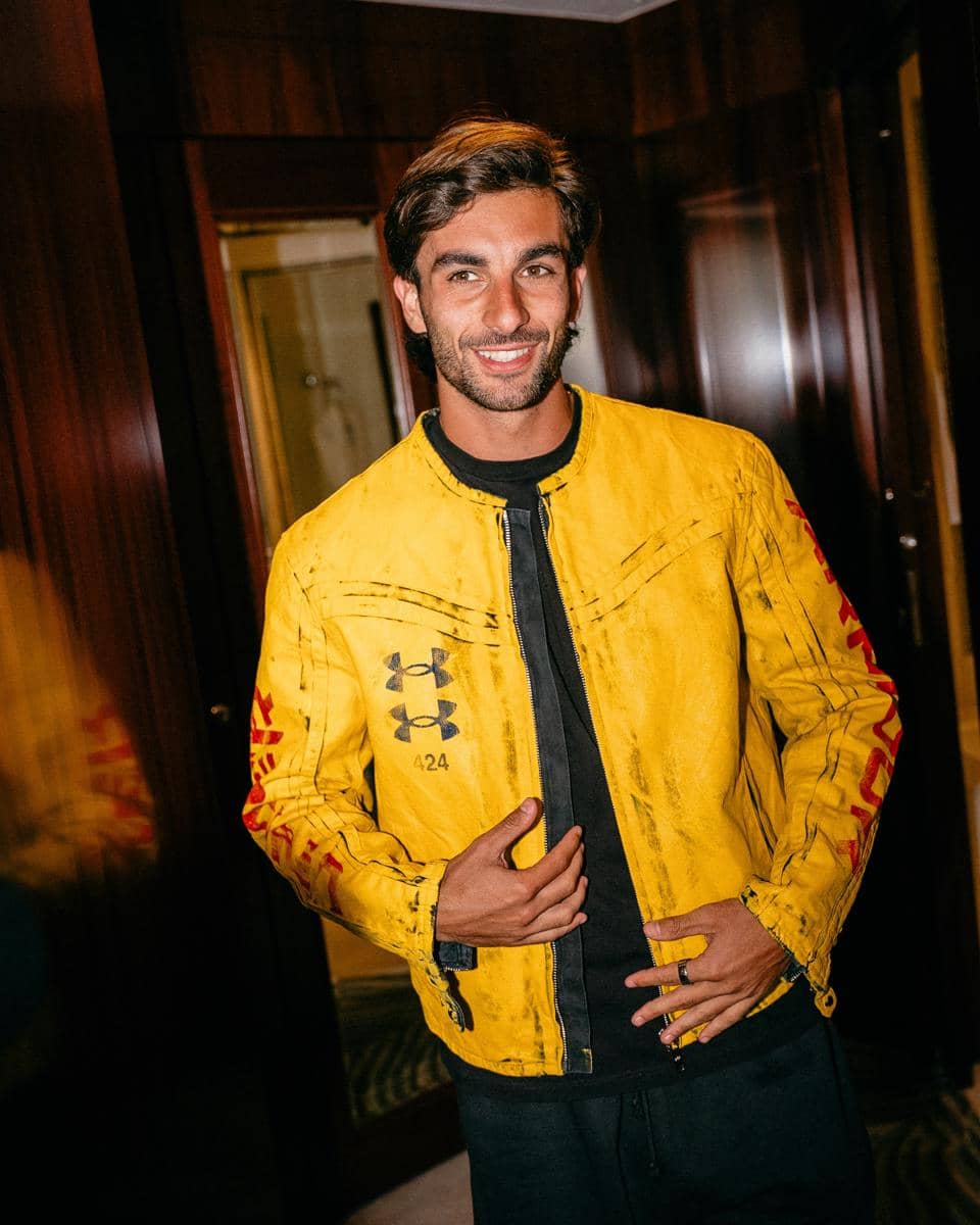 Seorang pria tersenyum mengenakan jaket kuning dengan logo Under Armour di dalam ruangan bernuansa kayu gelap.