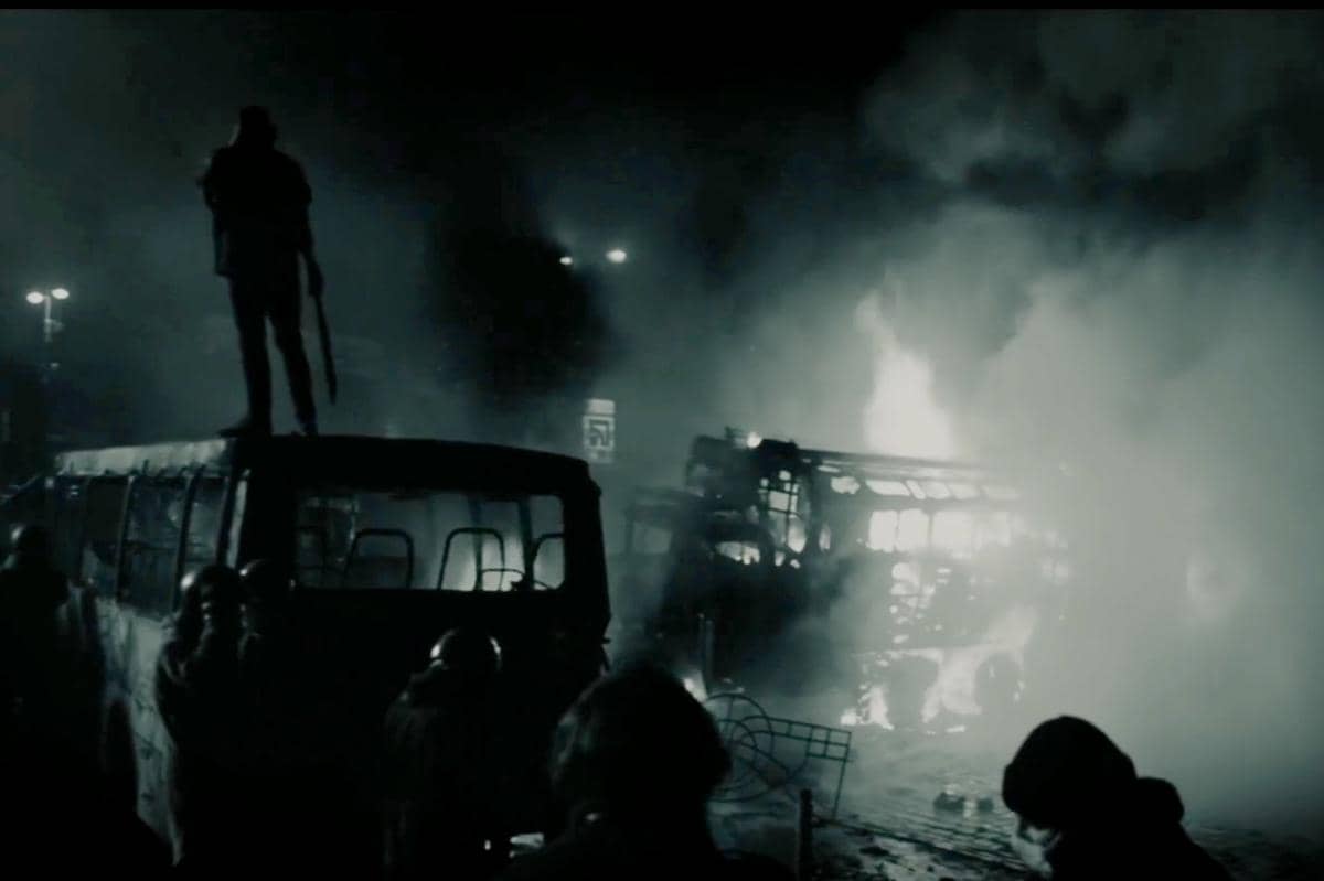 Adegan malam penuh asap dengan bus terbakar dan seseorang berdiri di atas kendaraan dalam cuplikan drama A Shop for Killers 2.